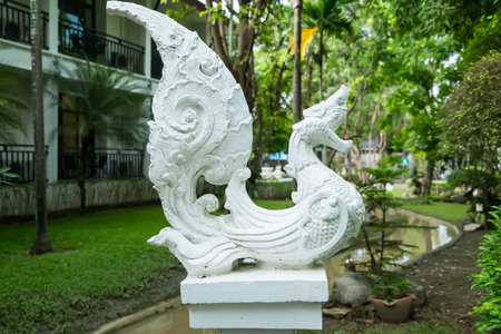 Ornate white stone sculpture of mythical bird with foliage backdrop in a lush garden settingの写真素材