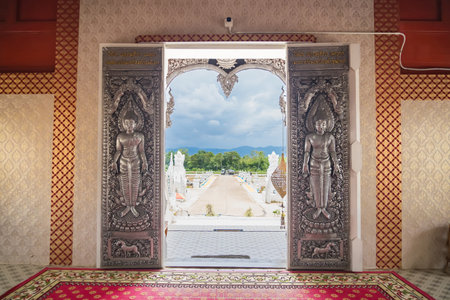 Chiang Mai, Thailand - July, 18, 2025 : View of scenic landscape through ornate open temple doors with intricate carvings in a traditional Thai style at Chiang Mai, Thailand.の写真素材
