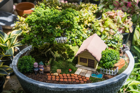 Miniature garden landscape in a pot featuring a small house, tree, figurines, and decorative pathway surrounded by greenery and plantsの写真素材