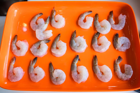 Raw peeled shrimp with tails arranged neatly on a bright orange tray in preparation for cooking or seafood dish preparationの写真素材