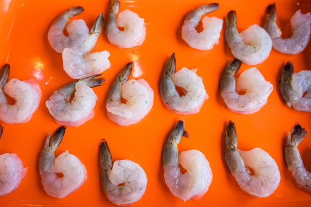 Raw peeled shrimp with tails arranged neatly on a bright orange tray in preparation for cooking or seafood dish preparationの写真素材