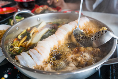 Close-up of a hot pot dish with shrimp, vegetables, and meat boiling in broth at an Asian restaurant with diners in the backgroundの写真素材