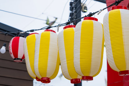 Row of yellow and white Japanese paper lanterns hanging outdoors in a festive urban street settingの写真素材