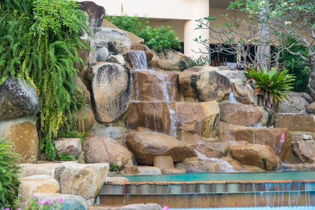 Chonburi, Thailand - November, 11, 2025 :Artificial rock waterfall feature in a landscaped garden with tropical plants and a pool in a resort settingの写真素材
