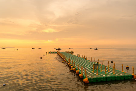 Floating dock extending into calm sea during golden sunrise with fishing boats on the horizon under a colorful skyの写真素材