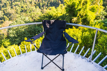 Black foldable camping chair placed on a circular balcony overlooking a lush green forest in bright...