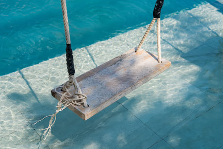 The serene scene of a wooden swing suspended above a crystal-clear swimming pool, inviting...