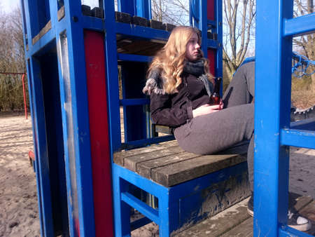 Teenage girl with sad look is sitting with a beer bottle on a climbing frame of a playgroundの写真素材