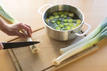 Hand holds leek for cutting with a knife. Sliced leek in pot with water next to it.の写真素材