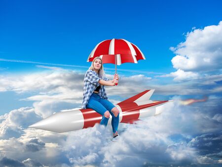 young woman sitting calmly on a rocket with an umbrella in her hand and flying confidently through the cloudsの写真素材