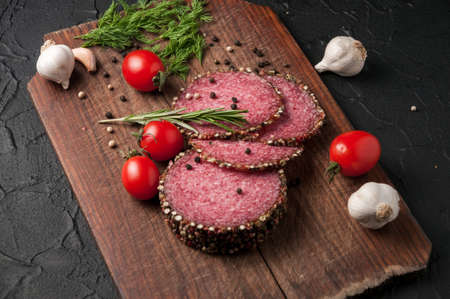 Smoked salami on a black table and a brown board. Sausages with rosemary, garlic and pepper and tomato cherry, dill, parsleyの写真素材