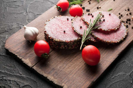 Smoked salami on a black table and a brown board. Sausages with rosemary, garlic and pepper and tomato cherry, dill, parsleyの写真素材