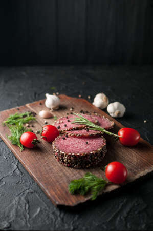Smoked salami on a black table and a brown board. Sausages with rosemary, garlic and pepper and tomato cherry, dill, parsleyの写真素材