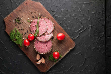 Smoked salami on a black table and a brown board. Sausages with rosemary, garlic and pepper and tomato cherry, dill, parsleyの写真素材