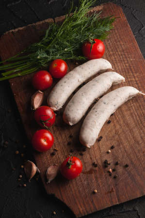 Three raw sausages on a wooden brown board on a black background with cherry tomatoes, parsley and dill, pepper and garlicの写真素材
