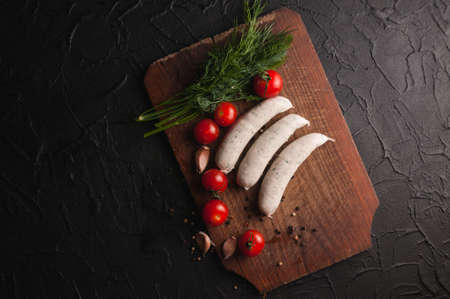 Three raw sausages on a wooden brown board on a black background with cherry tomatoes, parsley and dill, pepper and garlicの写真素材