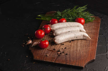Three raw sausages on a wooden brown board on a black background with cherry tomatoes, parsley and dill, pepper and garlicの写真素材