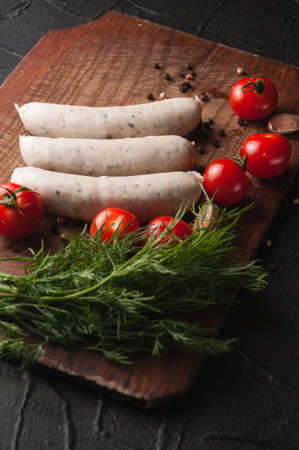 Three raw sausages on a wooden brown board on a black background with cherry tomatoes, parsley and dill, pepper and garlicの写真素材