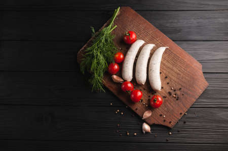 Three raw sausages on a wooden brown board on a black background with cherry tomatoes, parsley and dill, pepper and garlicの写真素材