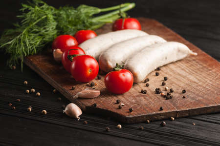 Three raw sausages on a wooden brown board on a black background with cherry tomatoes, parsley and dill, pepper and garlicの写真素材