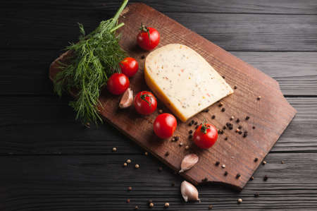 Cheese on a brown wooden board on a black wooden background, cherry tomatoes, pepper, greens, spices, dill, parsley. View from aboveの写真素材
