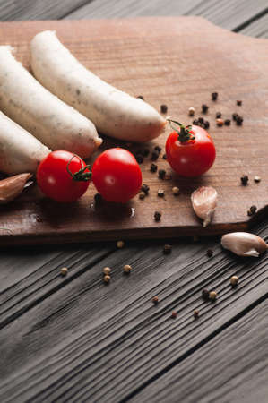 Three raw sausages on a wooden brown board on a black background with cherry tomatoes, parsley and dill, pepper and garlicの写真素材