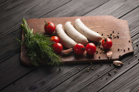 Three raw sausages on a wooden brown board on a black background with cherry tomatoes, parsley and dill, pepper and garlicの写真素材