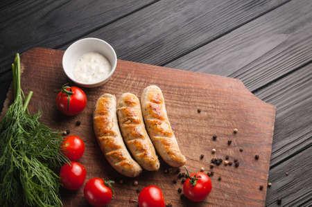 Grilled sausages grilled on a brown wooden board on a wooden background, six cherry tomatoes, greens, parsley dill, pepper, sauce. Place under the text and logo. Top view. Flat layの写真素材