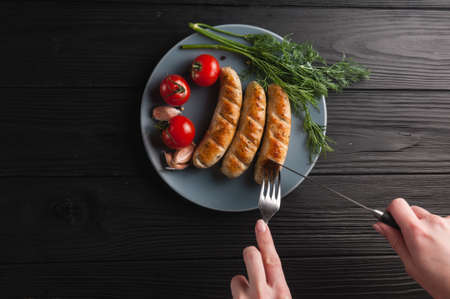 Three grilled sausages on a gray plate on a wooden black background lie also a cherry tomato, greens, garlic, knife, fork. View from above.の写真素材