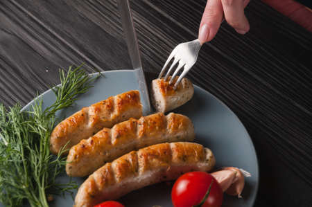 Three grilled sausages on a gray plate on a wooden black background lie also a cherry tomato,...