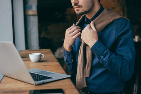 A man in a blue shirt sits in a cafe behind a laptop with a cup of coffee, straightens a brown sweater around his neck. Dark background, concept, without faceの写真素材