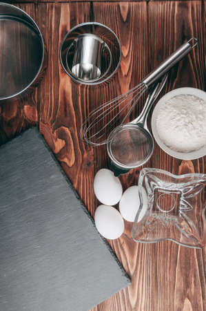 Ingredients in the dishes on a wooden brown table, where as well lie the whisk is full for flour, three chicken eggs and a slate board. View from above. Place under the textの写真素材