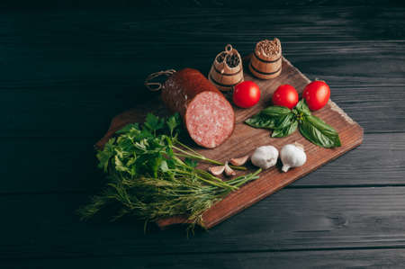 Smoked salami on a black table and a brown board. Sausages with rosemary, garlic and pepper and tomato cherry, dill, parsleyの写真素材