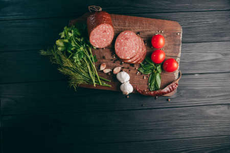 Smoked salami on a black table and a brown board. Sausages with rosemary, garlic and pepper and tomato cherry, dill, parsleyの写真素材