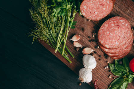 Smoked salami on a black table and a brown board. Sausages with rosemary, garlic and pepper and tomato cherry, dill, parsleyの写真素材