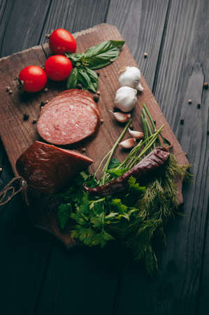 Smoked salami on a black table and a brown board. Sausages with rosemary, garlic and pepper and tomato cherry, dill, parsleyの写真素材