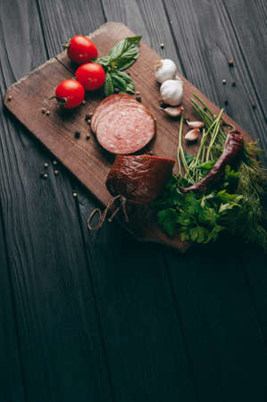 Smoked salami on a black table and a brown board. Sausages with rosemary, garlic and pepper and tomato cherry, dill, parsleyの写真素材