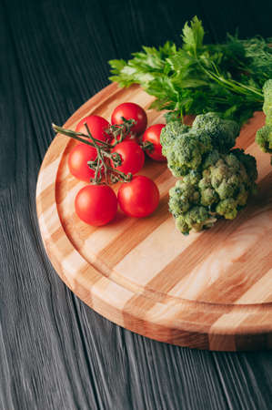 On a dark wooden table, on a round wooden board fresh green broccoli, parsley, dill and cherry tomatoes for your health. Recipe. Ingredients. Dietary food. Place under the text. View from aboveの写真素材