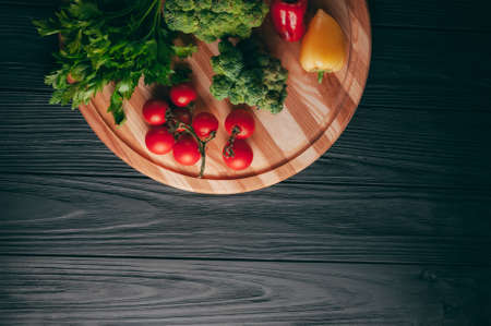 Fresh vegetables: cherry tomatoes, broccoli, pepper, dill, parsley on a round wooden board, on a dark wooden table. Recipe. Ingredients. Dietary food. Place under the text. View from aboveの写真素材