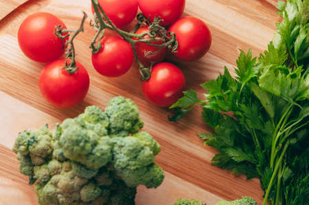 On a dark wooden table, on a round wooden board fresh green broccoli, parsley, dill and cherry...