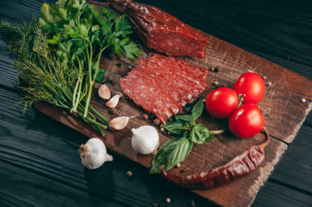 Smoked salami on a black table and a brown board. Sausages with rosemary, garlic and pepper and tomato cherry, dill, parsleyの写真素材