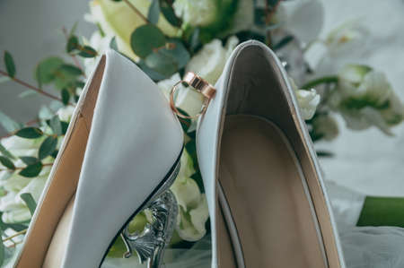 Wedding white bride shoes with a bouquet of white roses and other flowers, wedding rings on a stoolの写真素材