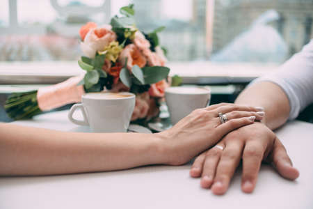 The bride and groom hold hands, holding expensive wedding rings with white gold, on the background of a wedding bouquet and two cups of coffee on a white table.の写真素材