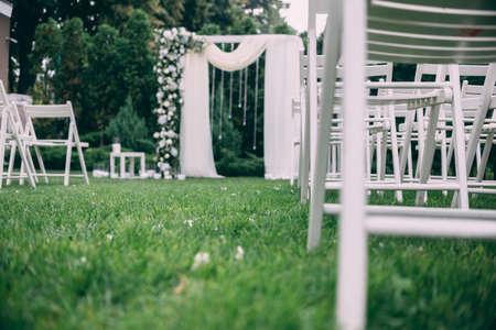 Beautiful wedding set up. Area of the wedding ceremony. Round arch, white chairs decorated with flowers, greenery. Cute, trendy rustic decor. Part of the festive decor, floral arrangement.の写真素材