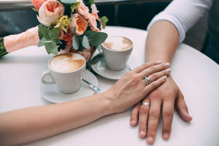 The bride and groom hold hands, holding expensive wedding rings with white gold, on the background of a wedding bouquet and two cups of coffee on a white table.の写真素材
