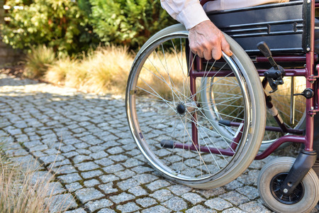 Close-up of old male hand on wheel of wheelchair during walk in parkの写真素材