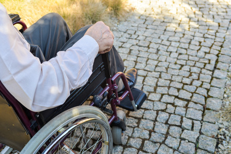 Close-up of old male hand on wheel of wheelchair during walk in parkの写真素材