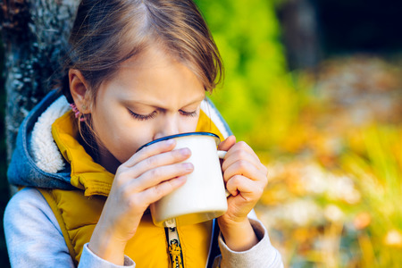 child girl drinking chocolate from a cup dressed in a warm yellow vest in autumn sceneryの写真素材