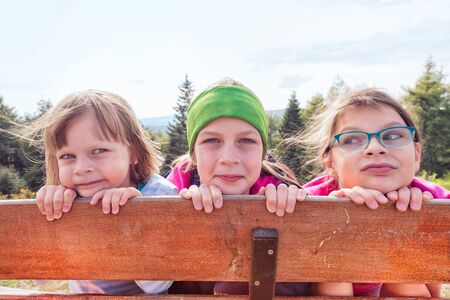 Three sisters on a mountain trip hiding behind a wooden fence - forest and mountains in the background behind themの写真素材