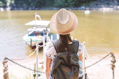 Girl in a straw hat and backpack on the banks of the river in anticipation of a cruise on a tourist boatの写真素材